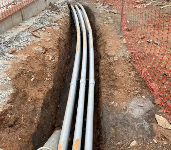 Bundled conduit exposed in an open trench behind orange safety fencing